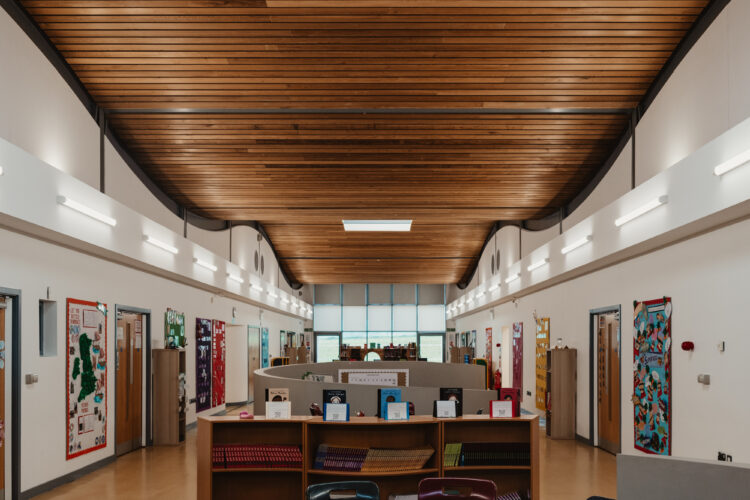 Image shows the internal of the hall within St James the Great Academy, a wooden curved ceiling with bright led lighting rods on either side.