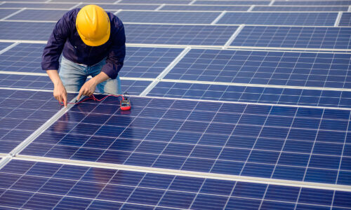Solar panels on a roof up close and a while male wearing a yellow hard hat on the solar panel, fixing it.
