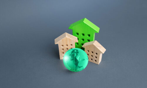 A dark grey background with three houses made of wood. Two are beige, one is painted bright green and in front of them sits a marble that looks like the world.