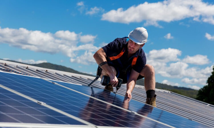 A white male workman wearing a white helmet, on top of roof top solar panels.