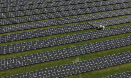 An aerial, drone view of Bowerhouse II solar park in Somerset. There are rows and rows of solar panels with a tractor along one line, cleaning the panels. The green grass can be seen between the rows.