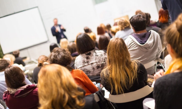 Taken from the back of a university lecture with the teacher in the background and slightly blurred. The students backs are towards the camera and are more in focus the more they come to the foreground.